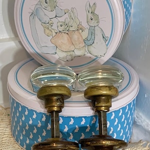 May include: Two vintage glass and brass door knobs. The knobs have a clear, rounded glass top and a brass base. The background includes two round tins with a blue bunny pattern and a Peter Rabbit illustration.