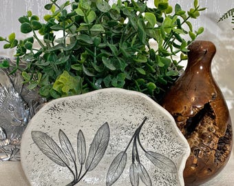 May include: A white, irregularly shaped ceramic dish with a black leaf design. A brown, wooden vase and green foliage are behind the dish. A silver bunny-shaped object is partially visible on the left.