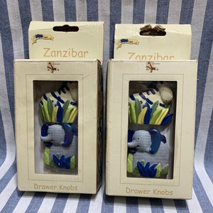 May include: Two packages of drawer knobs featuring a zebra and elephant design. The packages are white with blue and green accents and the text "Zanzibar" and "Drawer Knobs".