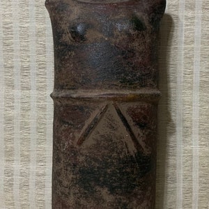 May include: A dark brown ceramic sculpture of a female figure with a simple, geometric design. The sculpture has a rounded, cylindrical body and a flat, circular head with a raised rim. The figure's arms are folded across its chest, and its legs are not visible.