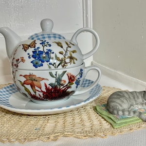 May include: A white porcelain tea set with floral and butterfly designs, including a teapot, teacup, and saucer. The lid has a blue and white checkered pattern. A gray cat figurine rests on a green pillow.