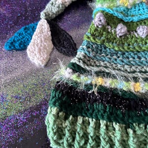 May include: A green, blue, and white crocheted hat with a long, striped tail. The hat has a black and white stripe pattern at the top and a green crocheted brim.