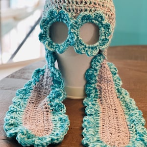 May include: A crocheted hat with eye holes and two long crocheted scarves attached. The hat is pink and green, and the scarves are blue and pink.
