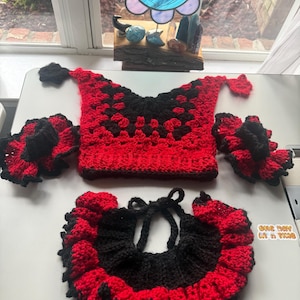 May include: A handmade crochet set featuring a red and black square-patterned top, two matching ruffled arm cuffs, and a ruffled collar with tie closures. The items are displayed on a white surface with a visible ruler measuring up to 14 inches.
