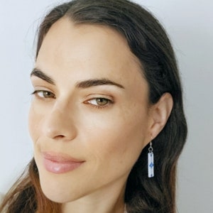 May include: A woman with long brown hair is wearing a pair of silver dangle earrings with a blue and white geometric design.