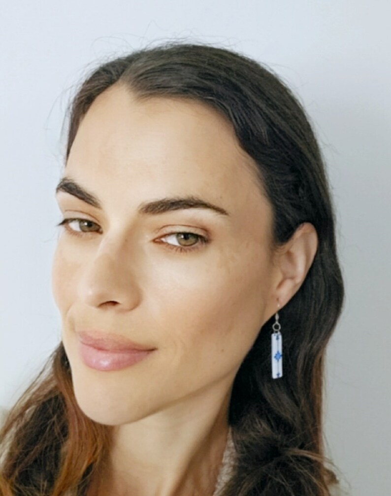 May include: A woman with long brown hair is wearing a pair of silver dangle earrings with a blue and white geometric design.
