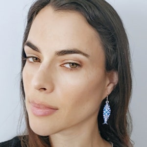 May include: A woman wearing a blue and white fish-shaped dangle earring. The earring is made of metal and has a blue and white enamel design.