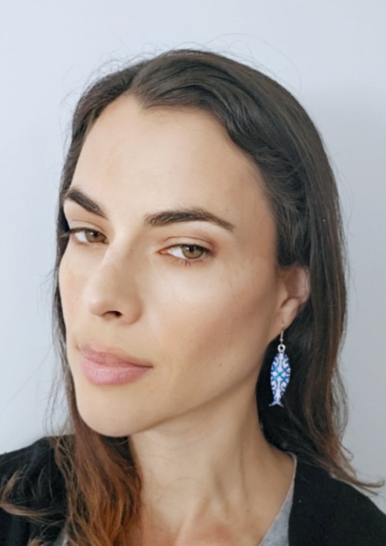May include: A woman wearing a blue and white fish-shaped dangle earring. The earring is made of metal and has a blue and white enamel design.