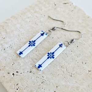 May include: A pair of dangle earrings with white ceramic rectangular charms featuring a blue floral pattern. The charms are attached to silver hooks.