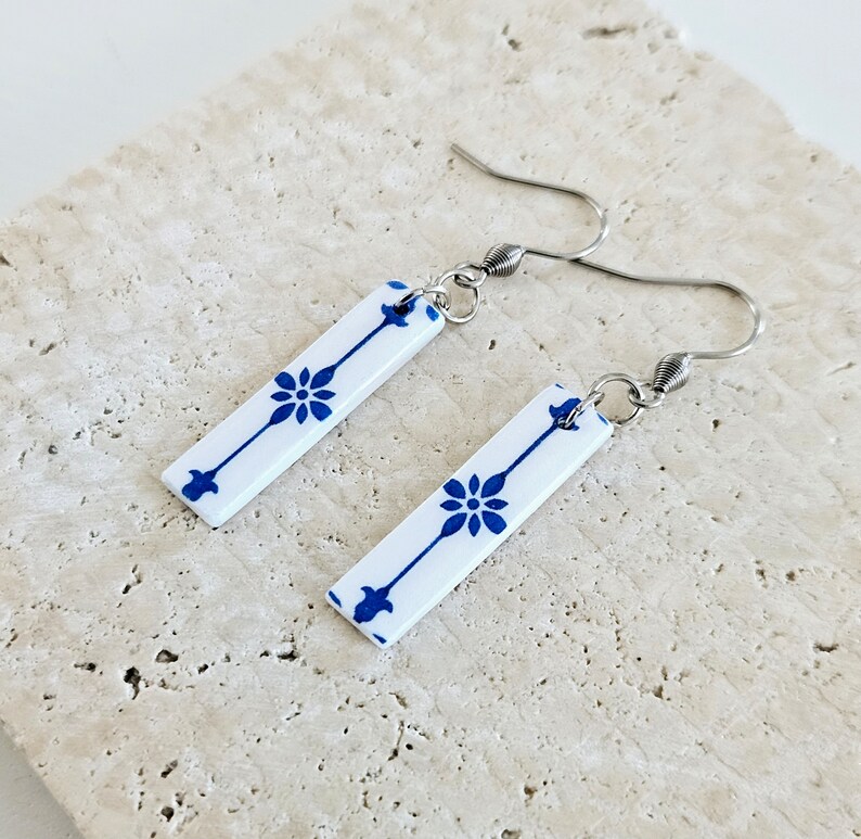 May include: A pair of dangle earrings with white ceramic rectangular charms featuring a blue floral pattern. The charms are attached to silver hooks.