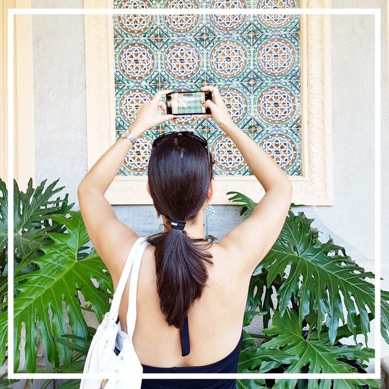 K&ouml;nnte beinhalten: Eine Frau mit langen braunen Haaren macht ein Foto einer gekachelten Wand mit einem Smartphone. Die Wand ist mit komplizierten geometrischen Mustern in Blau-, Gr&uuml;n- und Wei&szlig;t&ouml;nen verziert. Die Frau tr&auml;gt ein schwarzes Tanktop und eine wei&szlig;e Tasche.