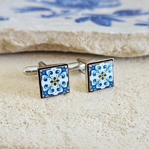 May include: A pair of silver cufflinks with blue and white tile-inspired designs. The tiles feature a yellow floral pattern.