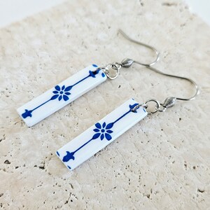 May include: A pair of white ceramic earrings with blue floral designs. The earrings are rectangular in shape and have silver hooks.