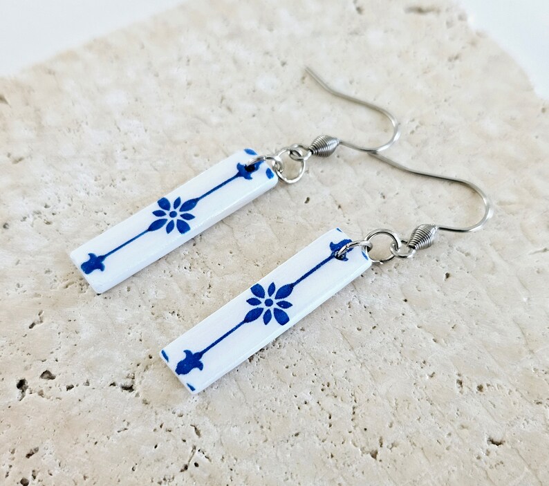 May include: A pair of white ceramic earrings with blue floral designs. The earrings are rectangular in shape and have silver hooks.