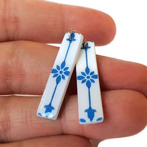 May include: A pair of white ceramic earrings with a blue floral design. The earrings are shaped like long rectangles with rounded edges.