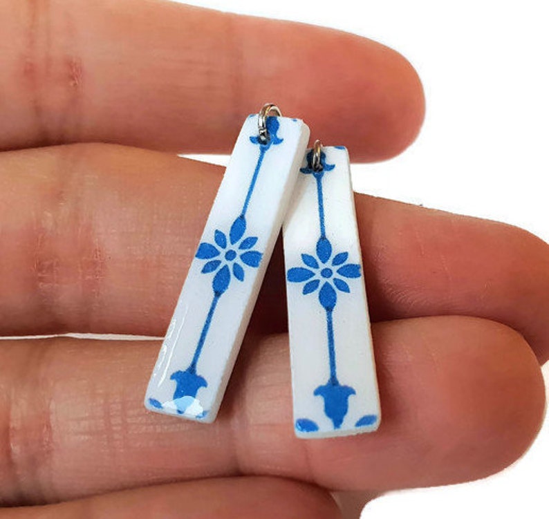 May include: A pair of white ceramic earrings with a blue floral design. The earrings are shaped like long rectangles with rounded edges.