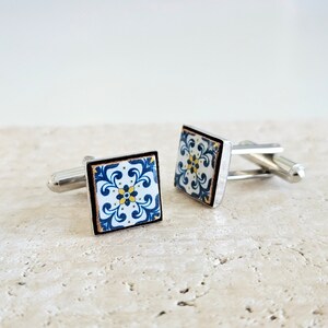 May include: A pair of silver cufflinks with blue and white patterned square tiles. The tiles have a floral design with yellow accents.