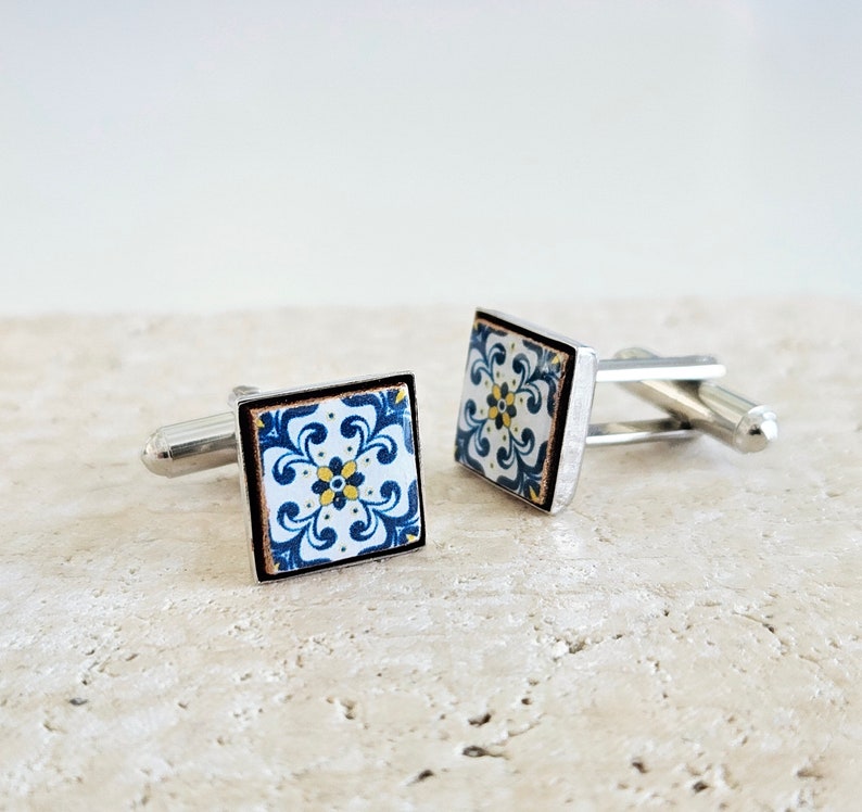 May include: A pair of silver cufflinks with blue and white patterned square tiles. The tiles have a floral design with yellow accents.