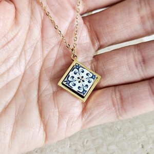 May include: A gold chain necklace with a square pendant featuring a blue and white tile design.