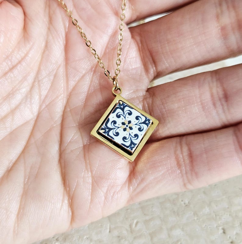 May include: A gold chain necklace with a square pendant featuring a blue and white tile design.