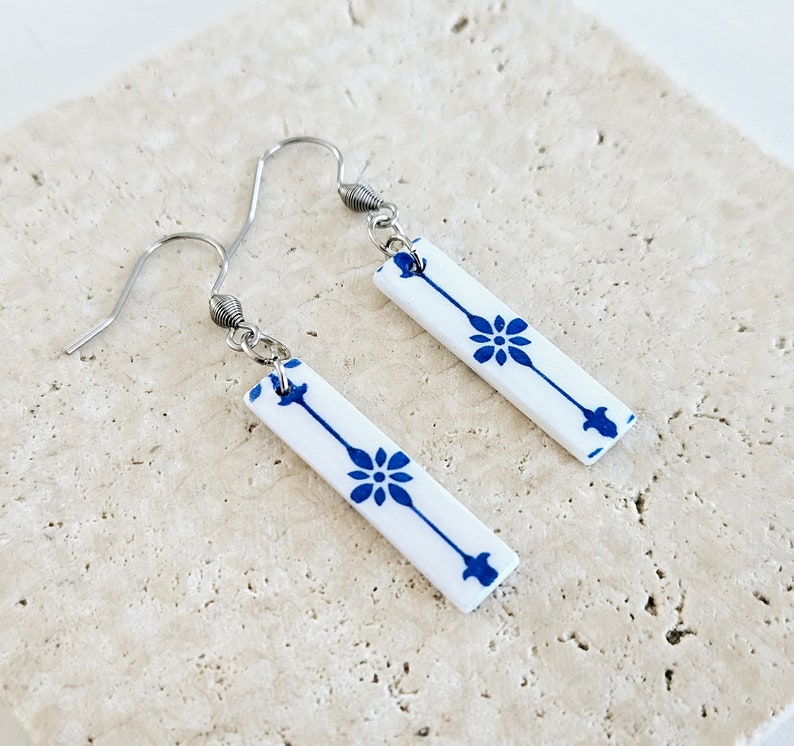 May include: A pair of silver dangle earrings with white ceramic rectangular charms. The charms have a blue and white floral pattern.