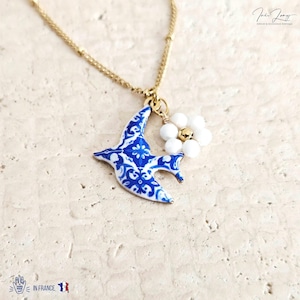 May include: A gold chain necklace with a pendant. The pendant features a blue and white patterned bird and a white flower with a gold centre. The necklace is displayed on a light beige surface. The words "IN FRANCE" are printed on the bottom left.