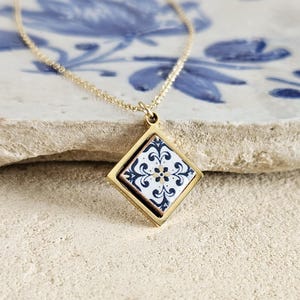 May include: A gold necklace with a square pendant featuring a white and blue patterned tile design.