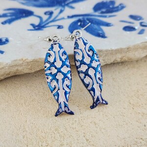 May include: A pair of blue and white fish-shaped earrings with a white filigree design. The earrings have silver hooks.