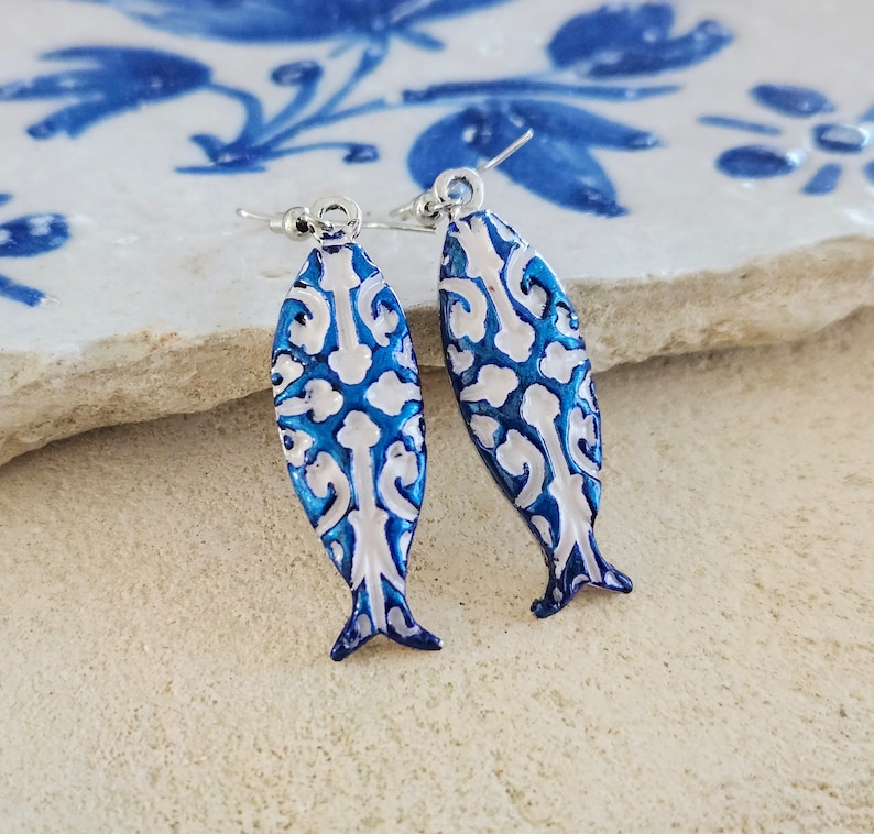 May include: A pair of blue and white fish-shaped earrings with a white filigree design. The earrings have silver hooks.