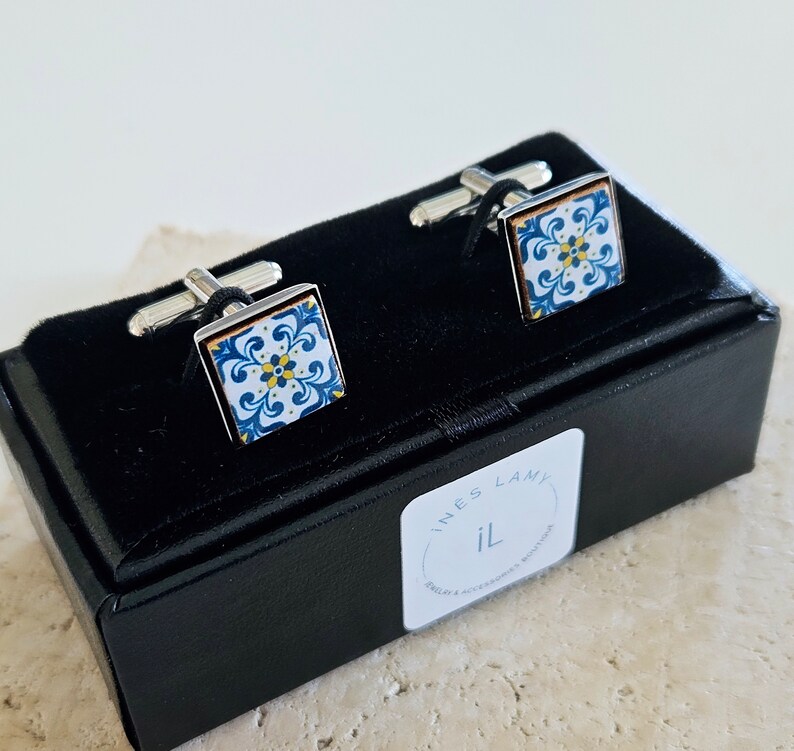 May include: A pair of cufflinks in a black box. The cufflinks feature a square design with a blue and white pattern. The box has a label that reads "Ines Lamy IL".