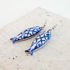 May include: A pair of silver fish-shaped earrings with a white and blue enamel design. The fish have a detailed floral pattern.