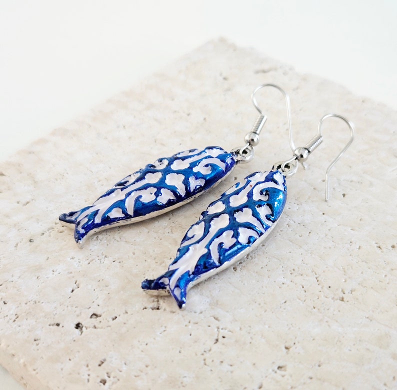 May include: A pair of silver fish-shaped earrings with a white and blue enamel design. The fish have a detailed floral pattern.