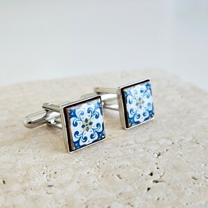 May include: A pair of silver cufflinks with blue and white patterned square tiles. The tiles have a floral design with yellow accents.