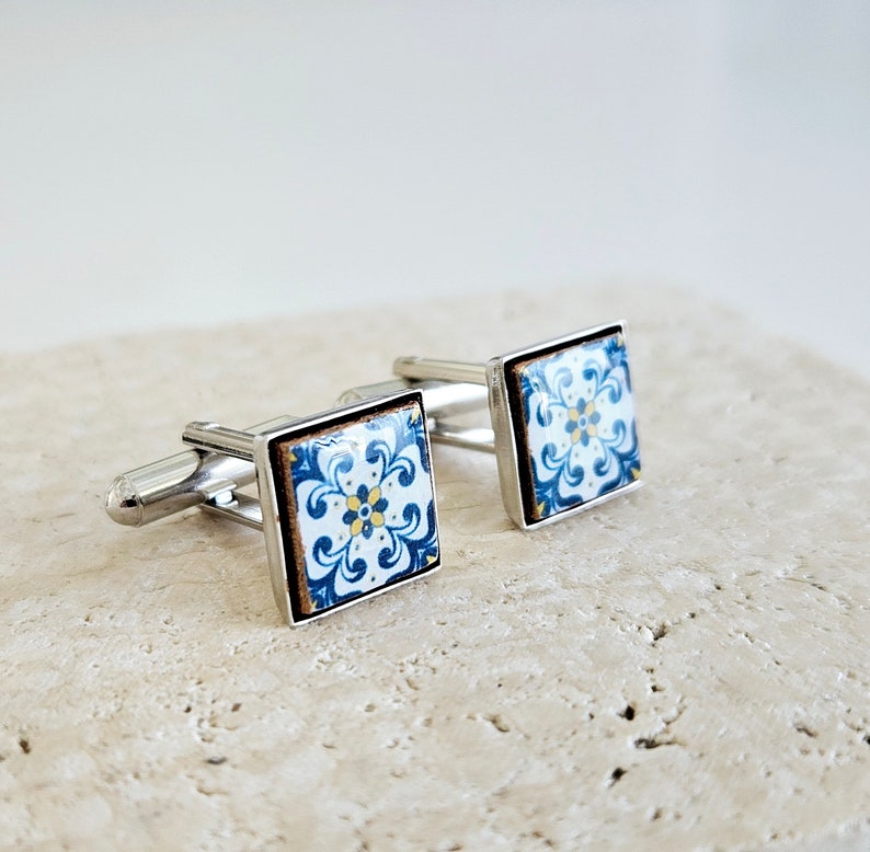 May include: A pair of silver cufflinks with blue and white patterned square tiles. The tiles have a floral design with yellow accents.