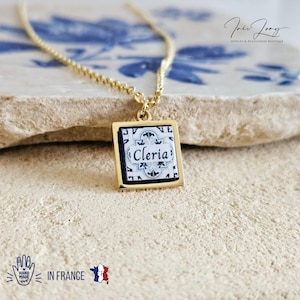 Peut inclure: Collier en chaîne dorée avec un pendentif carré orné d'un motif de carrelage blanc et noir avec le mot "Cleria" en lettres noires. Le pendentif est encadré d'or.