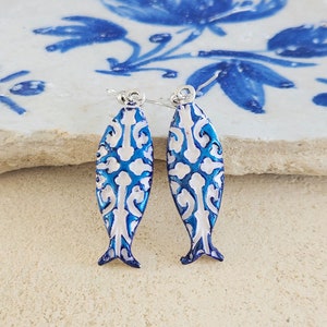 May include: A pair of blue and white fish-shaped earrings with a delicate white pattern. The earrings are made of metal and have a silver hook closure.