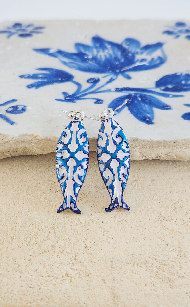 May include: A pair of blue and white fish-shaped earrings with a delicate white pattern. The earrings are made of metal and have a silver hook closure.