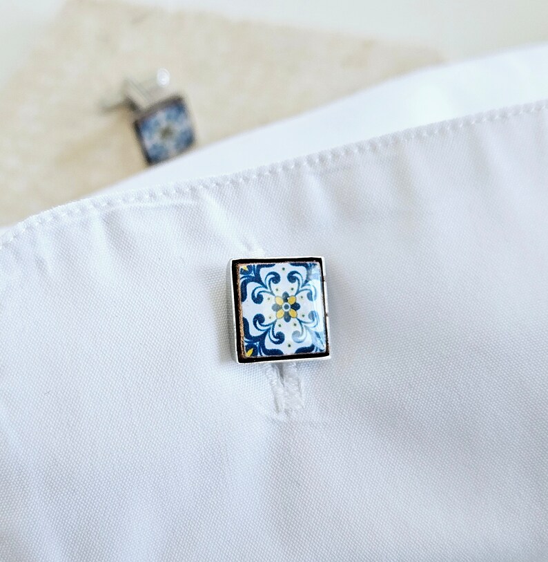 May include: A pair of cufflinks with a blue and white tile pattern. The cufflinks are silver and have a square shape.
