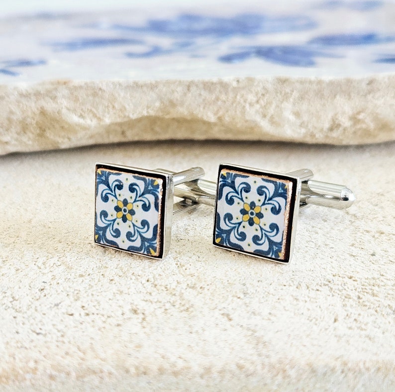 May include: A pair of silver cufflinks with a blue and white tile design. The tile features a yellow flower in the center.