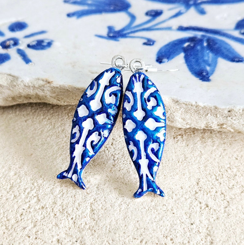 May include: A pair of blue and white fish-shaped earrings with a white filigree design. The earrings are made of polymer clay and have silver hooks.