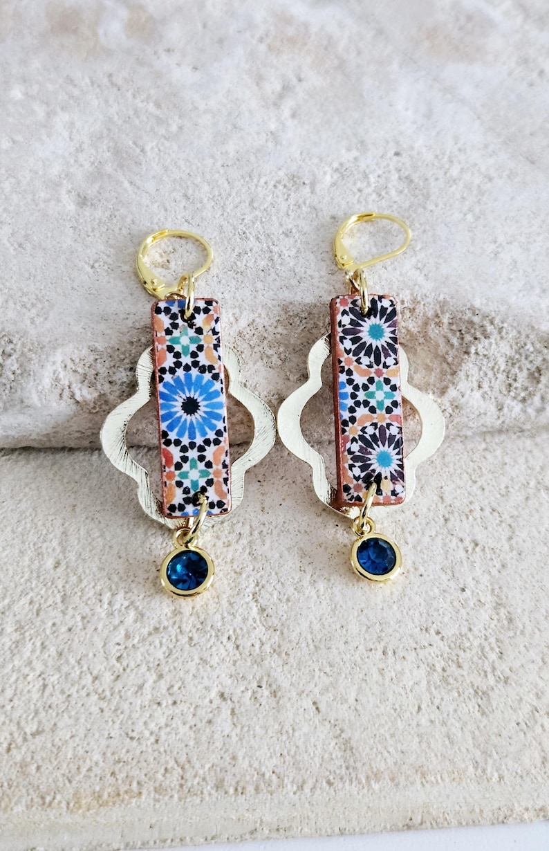 Peut inclure: Une paire de boucles d'oreilles en or avec un motif de carrelage bleu et blanc et une pierre pr&eacute;cieuse bleue suspendue. Les boucles d'oreilles ont un anneau en or et un motif de tr&egrave;fle &agrave; quatre feuilles en or.