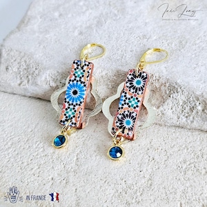 Peut inclure: Une paire de boucles d'oreilles en or avec un motif g&eacute;om&eacute;trique bleu et orange sur la partie pendante. Les boucles d'oreilles ont une petite pierre pr&eacute;cieuse bleue au bas.
