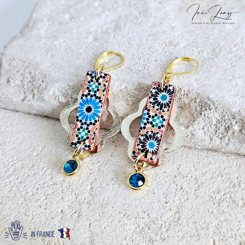 Peut inclure: Une paire de boucles d'oreilles en or avec un motif g&eacute;om&eacute;trique bleu et orange sur la partie pendante. Les boucles d'oreilles ont une petite pierre pr&eacute;cieuse bleue au bas.