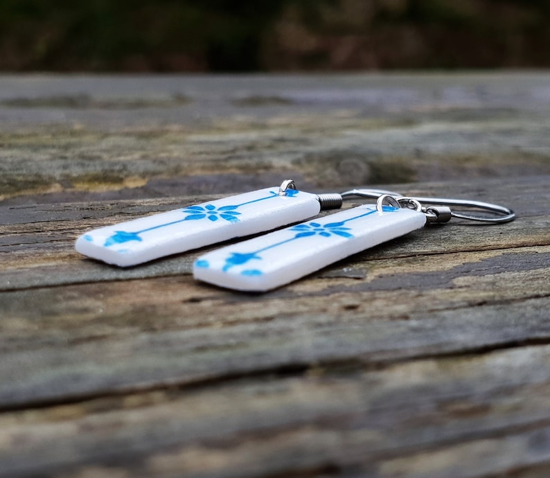 May include: A pair of white ceramic earrings with a blue floral pattern and silver hooks.