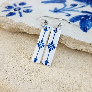 May include: A pair of white ceramic earrings with a blue floral pattern. The earrings are rectangular in shape and have a silver hook.