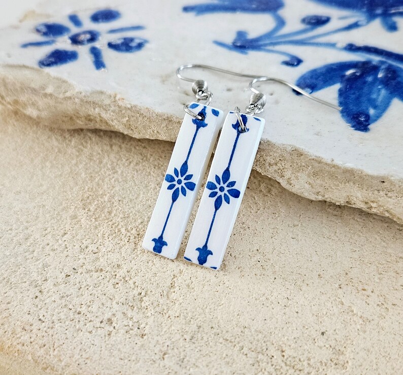 May include: A pair of white ceramic earrings with a blue floral pattern. The earrings are rectangular in shape and have a silver hook.