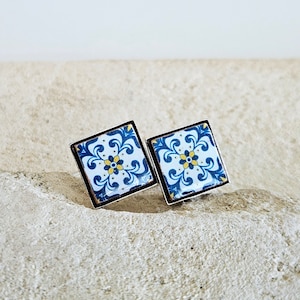 May include: A pair of square-shaped earrings with a blue and white tile pattern. The earrings have a silver metal frame.
