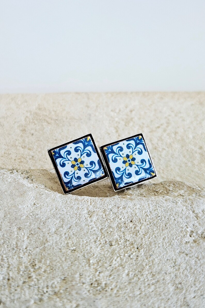 May include: A pair of square-shaped earrings with a blue and white tile pattern. The earrings have a silver metal frame.