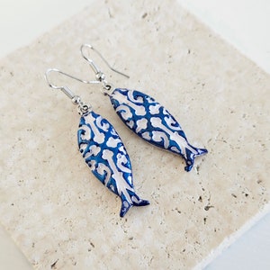May include: A pair of silver fish-shaped earrings with a blue and white patterned design.