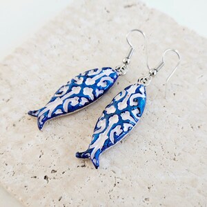 May include: A pair of blue and white fish-shaped earrings with a silver hook. The fish have a white filigree design on a blue background.